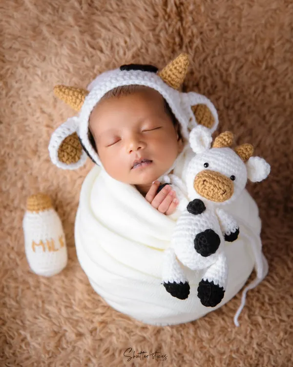 Holy cow, this is cute! A newborn dressed in a hand-knitted cow outfit, complete with a little milk bottle prop, sleeping on a soft brown blanket.