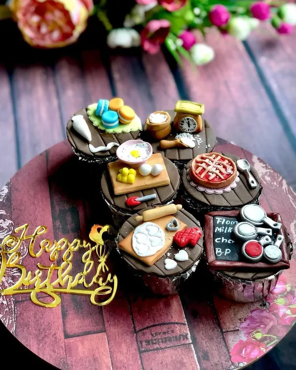 A perfect gift for a passionate baker. This set of customized cupcakes features miniature, handcrafted fondant toppers depicting a baker's life, including a tiny pie, macarons, a rolling pin, and a chalkboard with ingredients.