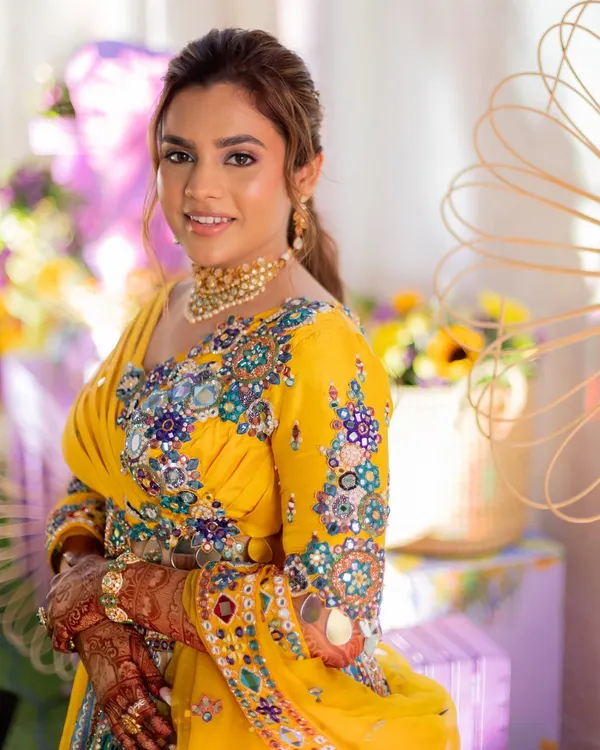 A colorful Haldi look for a vibrant bride. She is wearing a stunning yellow outfit with multi-colored embroidery, and her makeup is fresh and playful with a pop of color on the eyes.