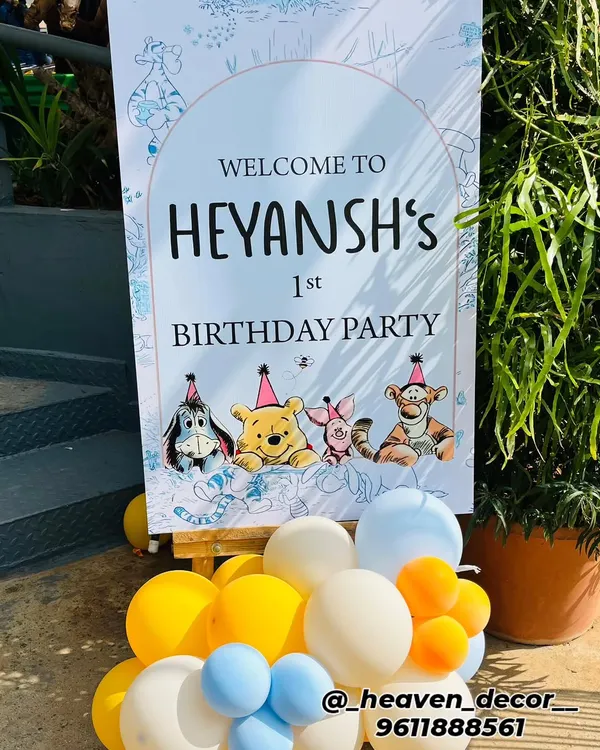 A charming welcome sign for a Winnie the Pooh first birthday party. The sign features characters from the Hundred Acre Wood and is displayed on an easel with a small balloon cluster.