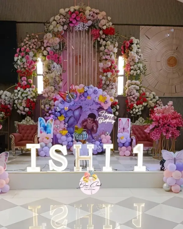 A grand stage decoration for a first birthday, featuring a massive floral arch and a butterfly-themed balloon setup with marquee letters.