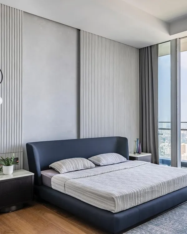 This minimalist master bedroom in 'The Grey House' overlooks the city and is designed for serenity. The feature wall combines smooth concrete texture with vertical fluting, creating a subtle play of light and shadow.