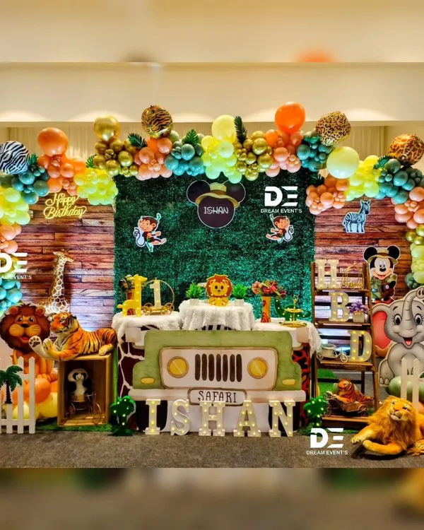 A full safari adventure stage for Ishan's first birthday. The setup includes a grass-green backdrop, a jeep-shaped table, and cutouts of all his favorite jungle animals like the lion, elephant, and monkey, all under a colorful balloon arch.