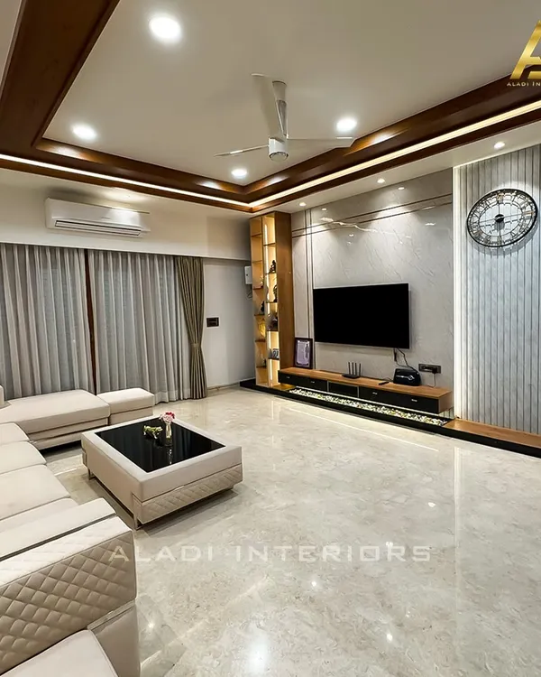 A spacious and modern living room designed for comfort and elegance. The space features a custom quilted L-shaped sofa, a sleek coffee table, and an elaborate TV unit with integrated lighting and a large statement clock, all set against polished marble flooring.