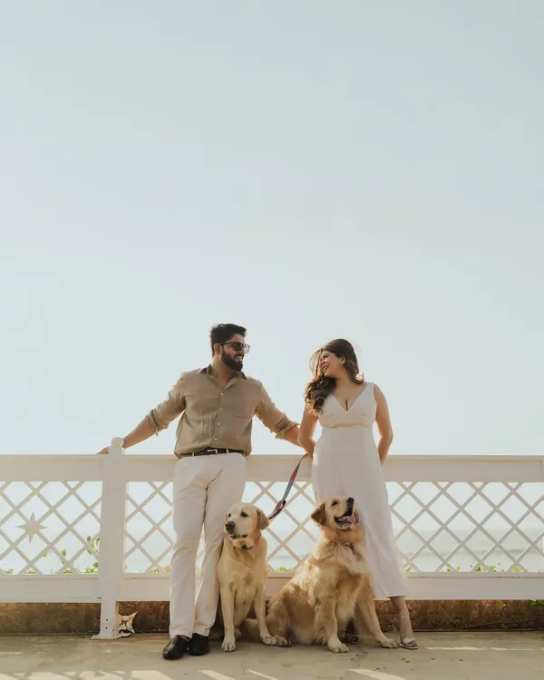 A beautiful family portrait of a couple with their two golden retrievers, posing by a white fence against a clear sky in Goa. This shot is a perfect example of a relaxed, pet-friendly pre-wedding shoot.