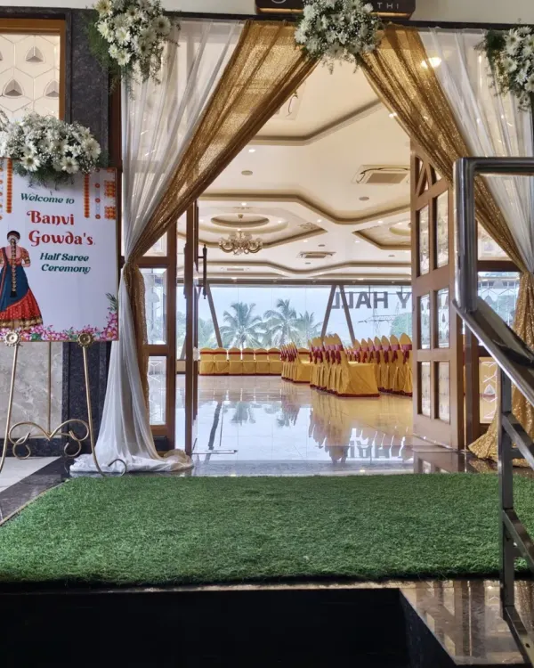A lovely entrance for a Half-Saree ceremony. The doorway is draped with elegant white and gold fabric, and a personalized welcome sign with a custom illustration greets the guests.