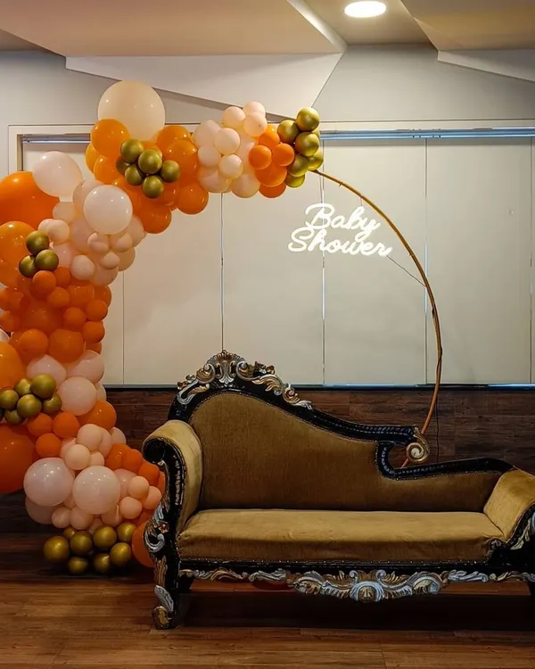 We create a special spot for the mom-to-be with this modern baby shower setup. It features a lush organic balloon arch in orange, white, and gold, a custom "Baby Shower" neon sign, and an elegant chaise lounge for comfort and perfect photos.