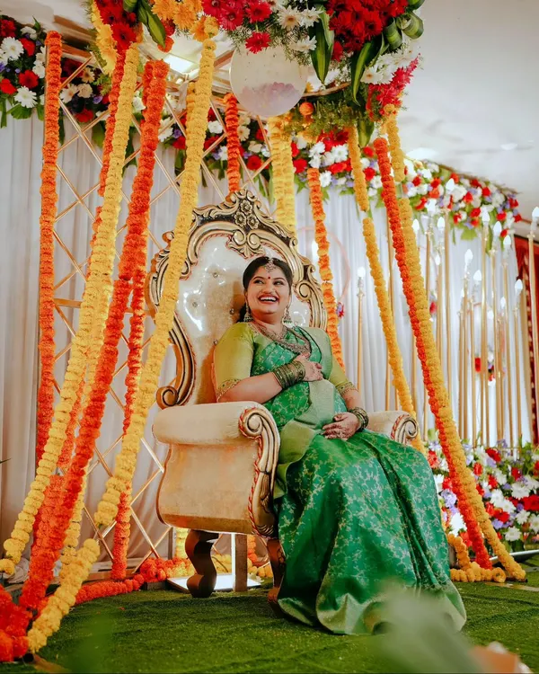 The mother-to-be looking radiant on an ornate throne during her Sreemantham ceremony. The vibrant floral decor with marigolds and roses creates a festive and traditional atmosphere.