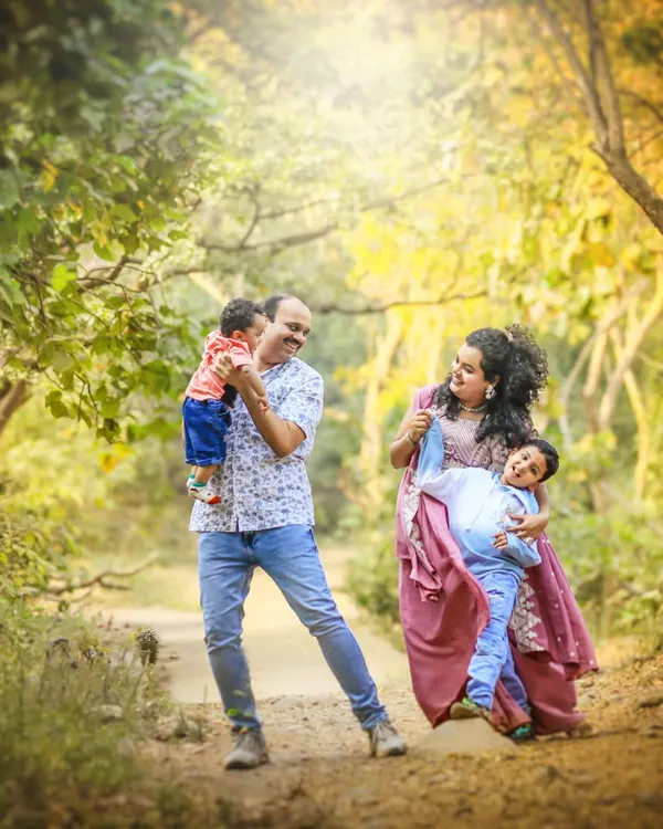 A family portrait alive with motion and joy. The father lifts the baby, the mother holds her older son, and the sunlight filtering through the trees adds a touch of magic to this candid moment.