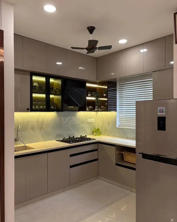 This L-shaped modular kitchen is designed for efficiency and style. It features handleless cabinets in a modern beige tone, a built-in hob, and under-cabinet lighting that illuminates the workspace. The black-framed glass cabinets add a touch of contrast and provide display storage.