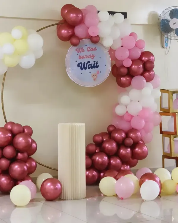 An elegant baby shower decoration in an apartment clubhouse. This setup features a ring balloon arch with pastel and chrome balloons, a custom "We Can Barely Wait" sign, and themed prop boxes.