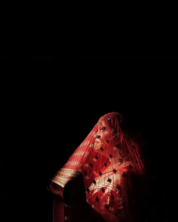 We use dramatic lighting to create powerful and artistic bridal portraits. This low-key photograph highlights the bride's silhouette and the rich texture of her red veil against a dark background, creating a truly memorable image.