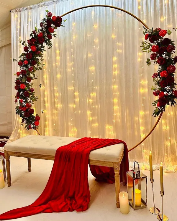 For a classic and passionate statement, you can’t go wrong with deep red roses. I designed this romantic backdrop with a full floral arch over a simple white bench, set against a curtain of warm fairy lights. This elegant stage decoration is perfect for making your engagement photos pop.