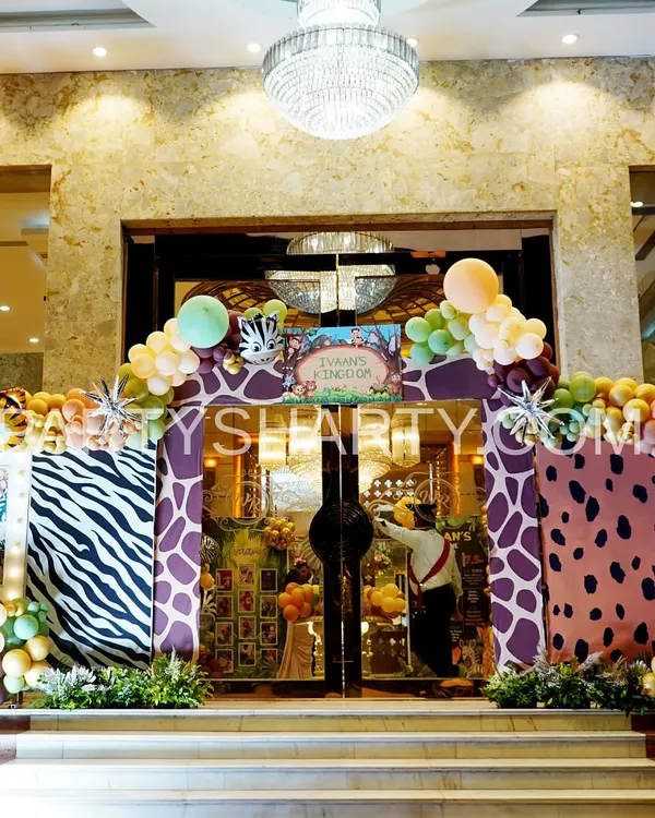 A grand jungle safari entrance featuring zebra and giraffe print arches, lush greenery, and a personalized 'Kingdom' sign for the birthday boy.