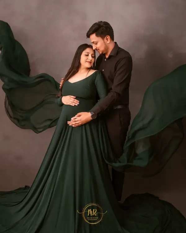 An intimate couple's portrait in our studio. The partner embraces the mother-to-be from behind, both gently holding the baby bump, surrounded by flowing green fabric.