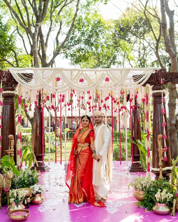 A beautiful couple framed by the grandeur of their wedding mandap. The decor is a blend of tradition and elegance, with rich wooden pillars, a ceiling of white floral strings, and accents of pink, creating a perfect backdrop for their special day.