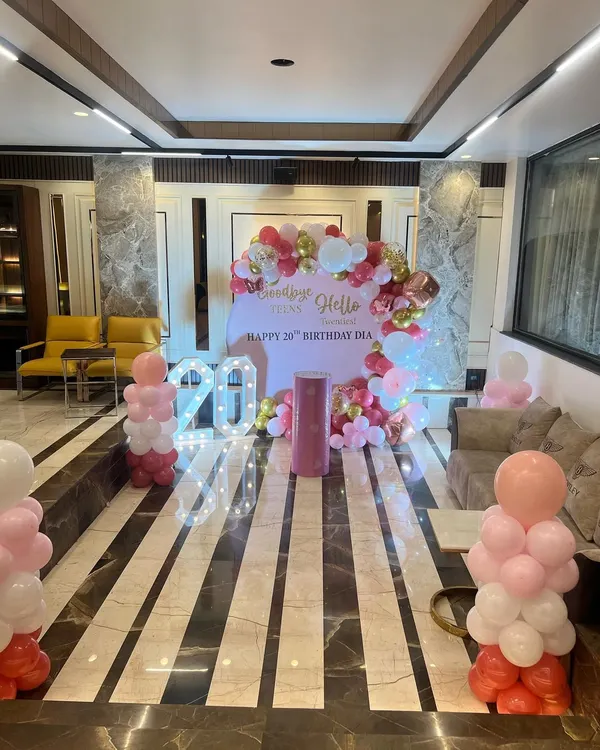 A wide shot of a 20th birthday decoration in a party hall, featuring a "Goodbye Teens, Hello Twenties" theme with pink, red, and gold balloons.