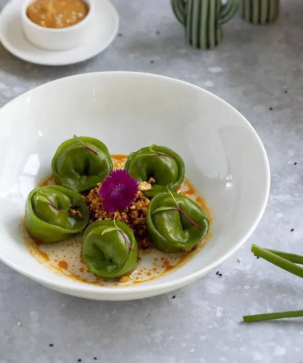 This is an example of modern, artistic food plating. The vibrant green of the spinach dumplings pops against the clean white bowl, while the careful placement of garnishes highlights the dish's elegance for a fine-dining menu.
