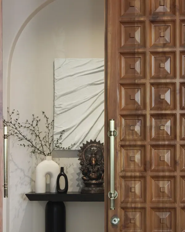 The entryway of the Jaipur House features a traditionally carved teakwood door. It opens to reveal a carefully composed scene with textile art and curated objects, setting a narrative tone for the entire home.