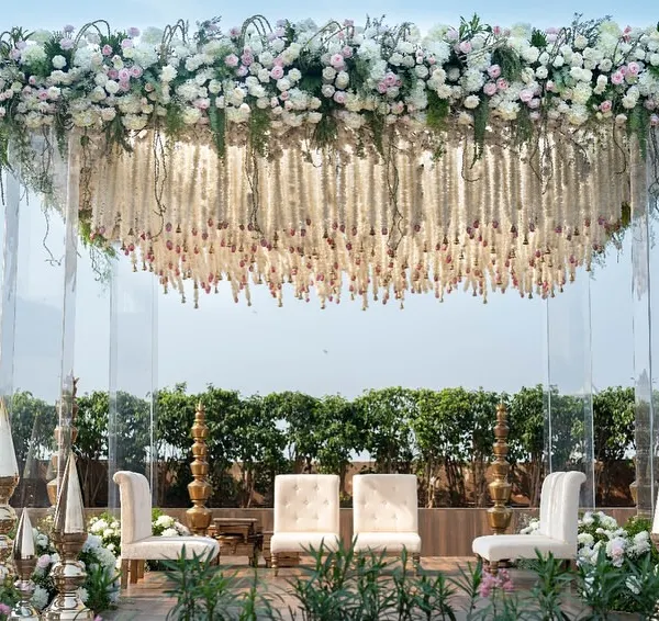 Our signature floating floral mandap, creating an ethereal atmosphere against the open sky. This design combines modern engineering with classic floral artistry, featuring a dense canopy of white and pink roses with cascading tuberose strings, making it a truly heavenly setting for exchanging vows.