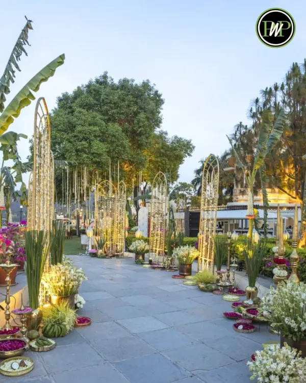 A truly grand welcome. This outdoor wedding entrance features ornate golden structures, lush banana plants, and an abundance of white and yellow flowers, creating a luxurious and traditional path for guests.