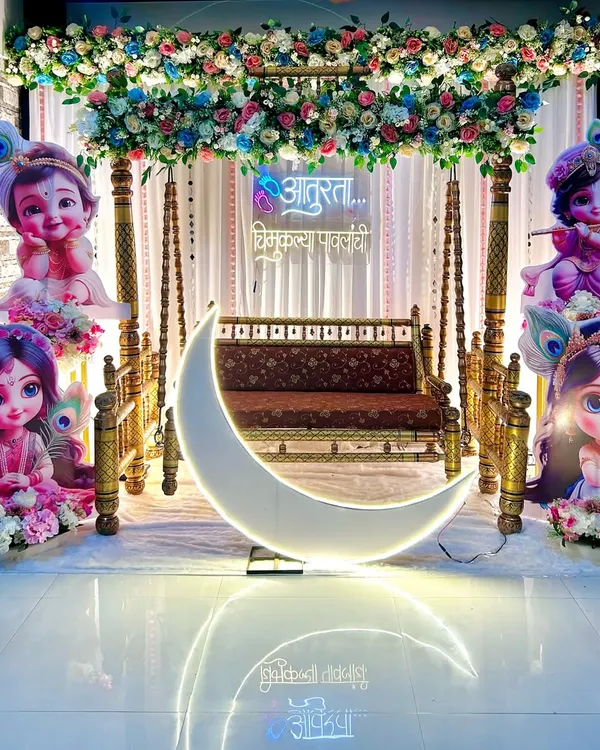 This 'Radha or Krishna' themed baby shower stage combines a traditional wooden swing and floral canopy with a modern LED moon prop. The neon sign in Marathi adds a personal touch to the backdrop.