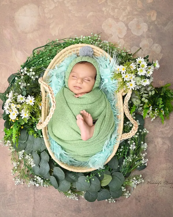 A perfect little bundle of peace. This newborn is swaddled in a soft green wrap, sleeping soundly in a basket surrounded by fresh greenery and daisies.