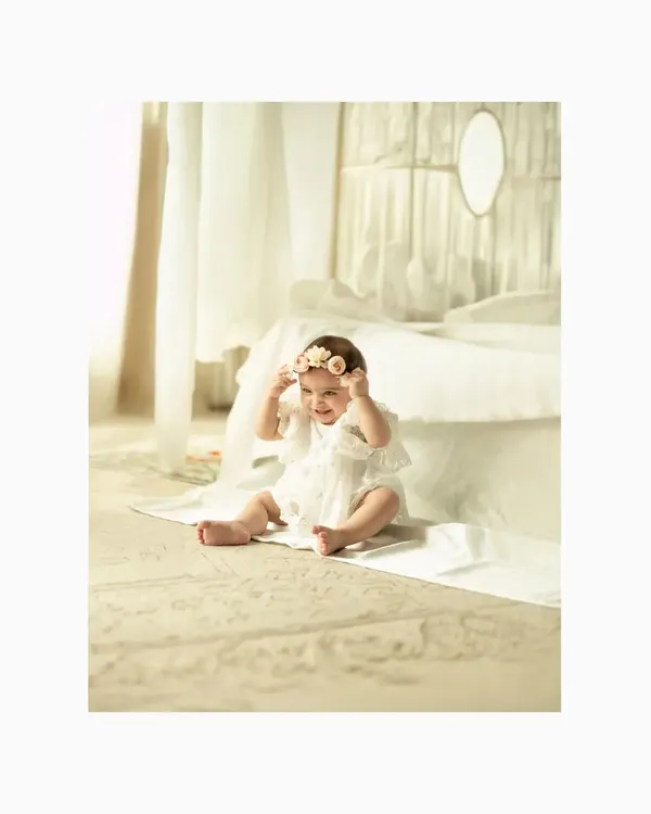 A happy baby girl sits on the floor in a bright white studio, playing with her floral headband. The clean, minimalist setting puts all the focus on her adorable expression and joyful spirit.