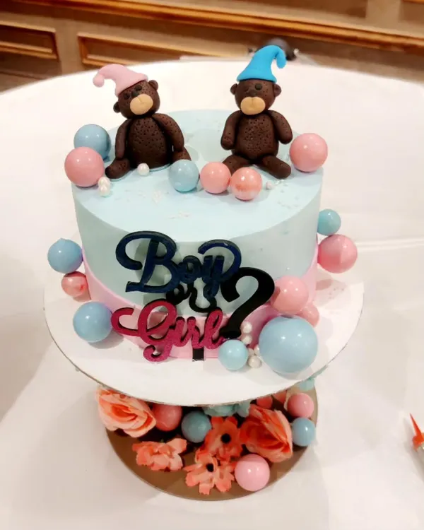 A beautiful two-tier cake for a "Boy or Girl?" gender reveal party. It features fondant teddy bears in pink and blue hats, surrounded by matching decorative balls.