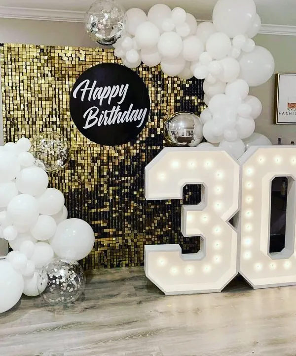 A chic and modern 30th birthday setup. The combination of a gold sequin wall, all-white balloon decor, disco balls, and bold marquee numbers creates a high-contrast, glamorous look perfect for a memorable party.