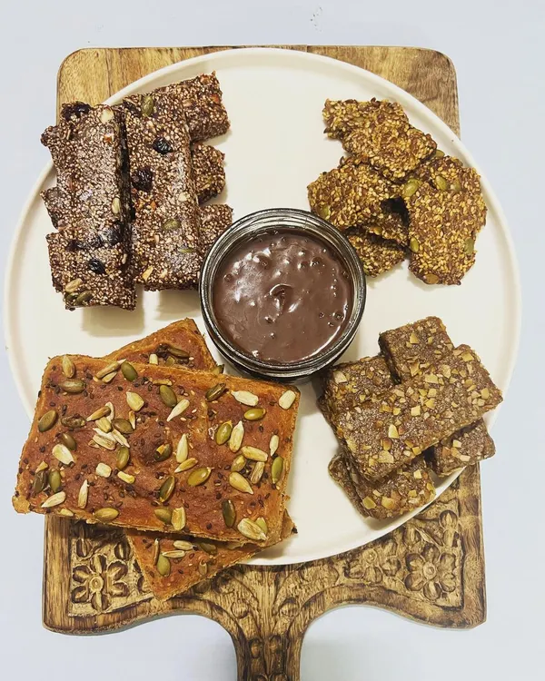 This platter showcases the heart of my kitchen. You'll find our signature flourless lentil bread, assorted protein-rich nutritional bars, crunchy seed crackers, and a jar of creamy, house-made hazelnut spread. Everything is 100% vegan and free from gluten, sugar, and preservatives.