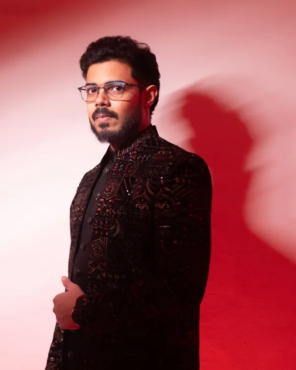 A stylish portrait of the groom-to-be against a red background. His direct gaze and sharp attire convey a sense of modern confidence.