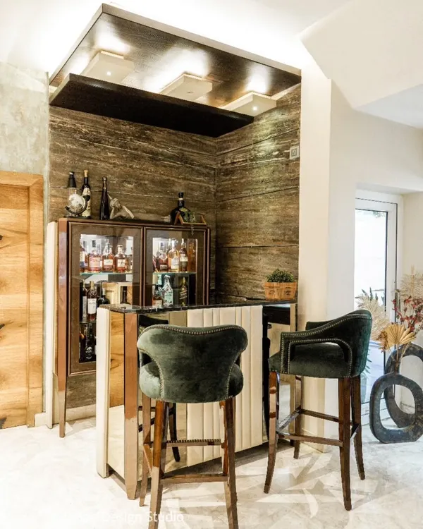 A luxurious home bar setup with Travertine marble walls, a dark wood ceiling, and plush forest-green velvet bar stools.