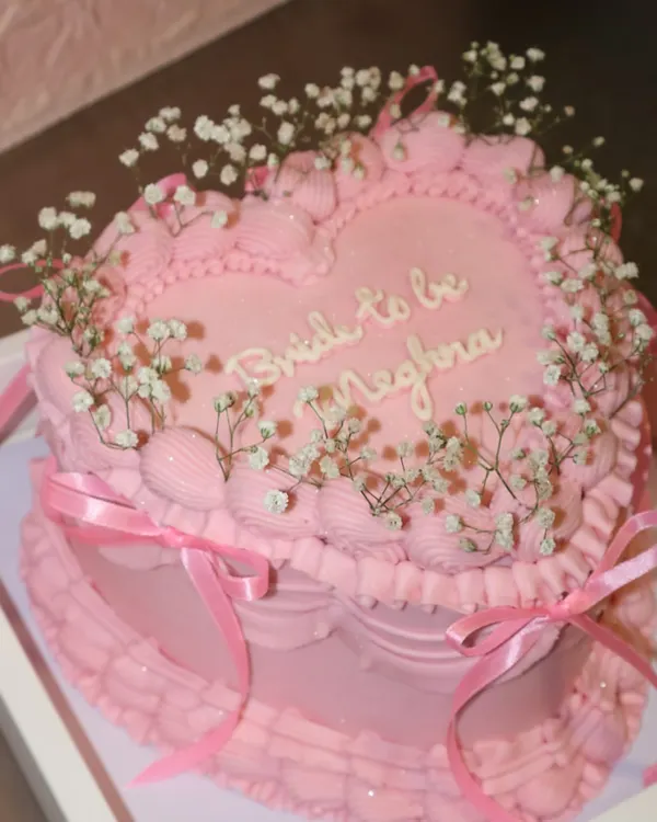A dreamy, heart-shaped bridal shower cake for a "Bride to be". This all-pink creation features intricate piping, delicate baby's breath flowers, and pretty satin ribbons for a truly romantic feel.