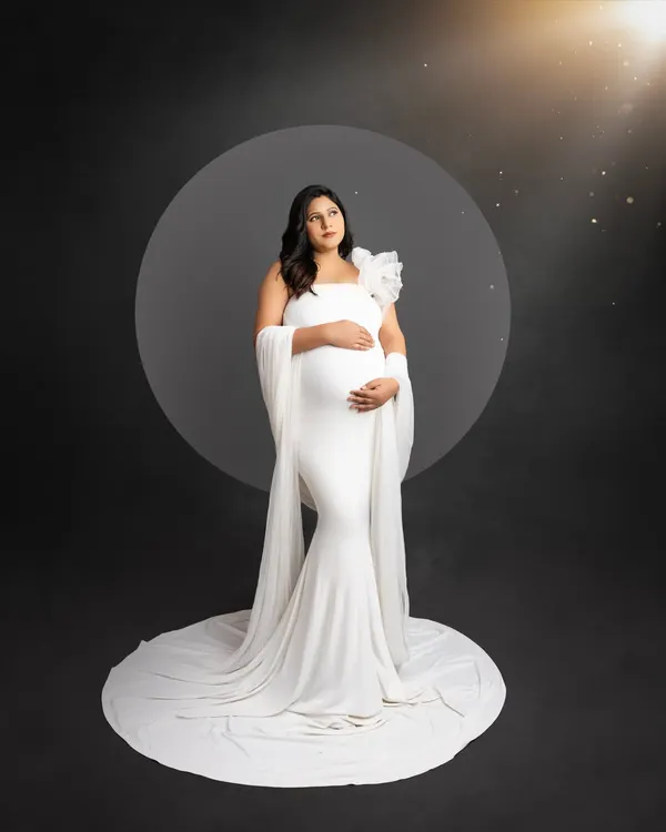 A classic and elegant solo maternity portrait. The mom-to-be wears a simple white gown, standing on a white circular platform against a dark background, with a soft light creating a celestial glow.
