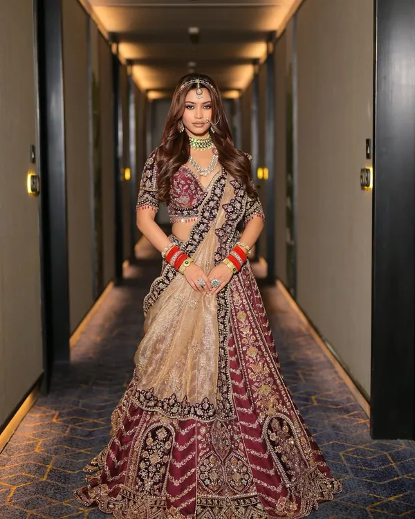 A full-length portrait of the modern maharani look, showcasing the deep maroon lehenga with intricate golden embroidery. The makeup is balanced to be impactful without overpowering the grandeur of the traditional attire.