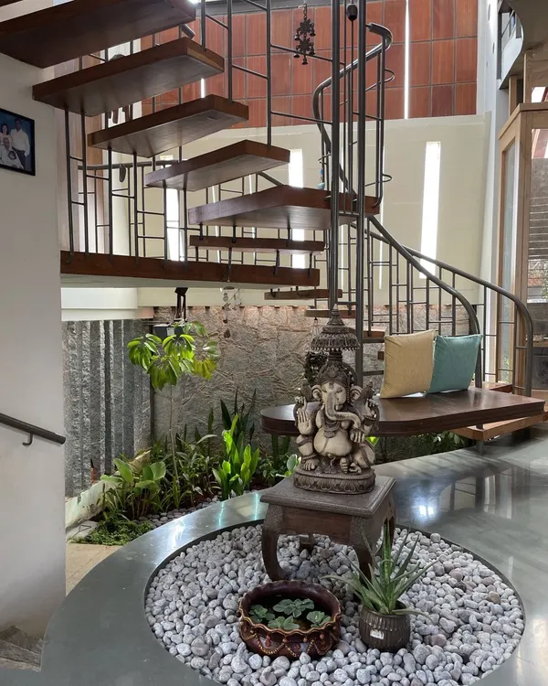 The entrance of a home should draw in positivity. Here, a spiral staircase wraps around a tranquil indoor courtyard. A Ganesha statue sits at the center, surrounded by white pebbles and plants, creating a serene and welcoming foyer that immediately connects the inside with nature.