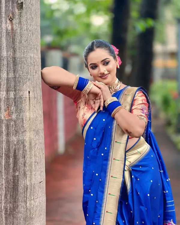 A beautiful outdoor shot of a model in a traditional blue Paithani saree. The makeup is kept classic with a prominent nath and choker, perfect for a pre-wedding photoshoot.