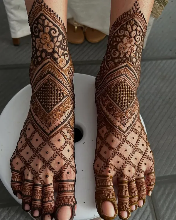 A stunning example of full-coverage feet mehendi. This design combines intricate floral bands around the ankle with a bold geometric diamond pattern that covers the entire foot.