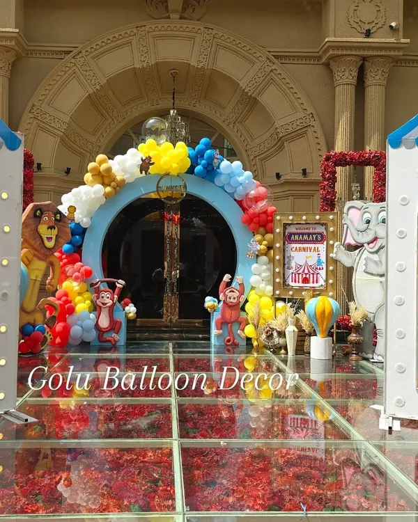 The entrance to a carnival-themed birthday party, featuring a colorful balloon arch and circus animal cutouts over a glass floor filled with flower petals.