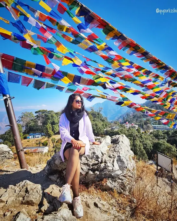 At the top of the world in Mussoorie, with Tibetan prayer flags fluttering in the wind. My travel styling focuses on layers, like this white jacket over a black dress, which are perfect for changing mountain weather.