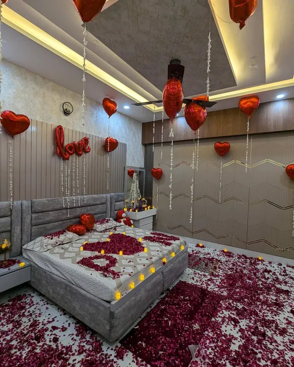 A classic romantic surprise setup. The room is filled with red heart balloons, a "LOVE" foil balloon, and a bed decorated with petals and LED candles spelling out "I ❤️ U".