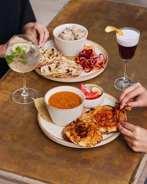 A table full of choices. Creamy chicken malai, rich dal, and flaky parathas paired with a gin and tonic and a deep red sour. This is what a meal at Comorin looks like.