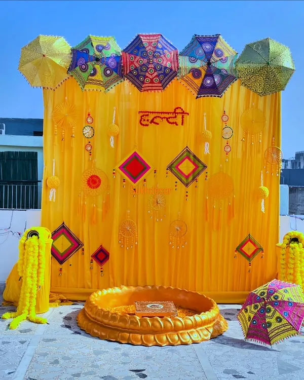 A vibrant and traditional Haldi ceremony setup on a rooftop. The decor is centered around a bright yellow backdrop, Rajasthani umbrellas, and marigold flower garlands.