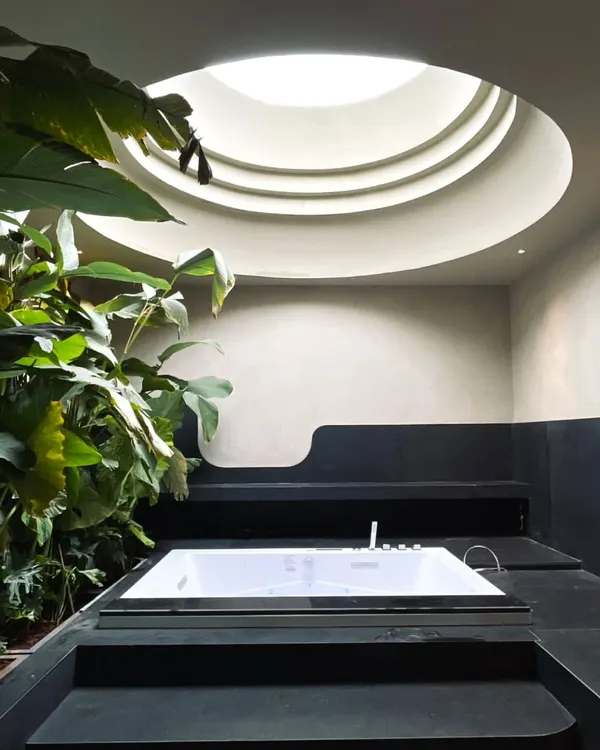 This contemporary bath space is designed around a central circular skylight, which washes the room in soft daylight. The whirlpool tub is positioned directly beneath, creating a focal point for luxury and tranquility, surrounded by lush indoor plants.