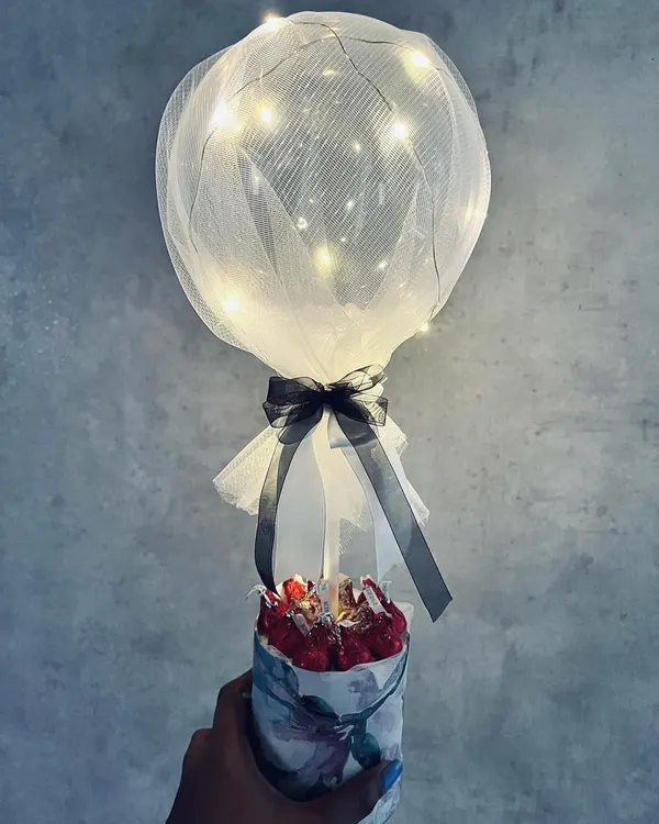 An enchanting balloon hamper featuring a transparent balloon with fairy lights wrapped in tulle, rising from a small floral box of chocolates. A magical gift for under ₹600.