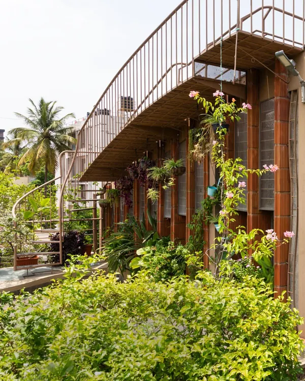 A spiral staircase connects the ground floor garden to the upper terrace, creating a continuous green pathway. The home is wrapped in plants, with creepers and hanging pots that blur the line between the building and the garden.