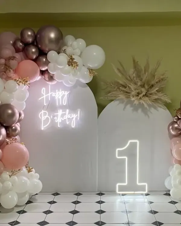 A chic and modern first birthday setup with white arched backdrops, a neon sign, a "1" marquee number, and a beautiful balloon arrangement in rose gold, pink, and white.