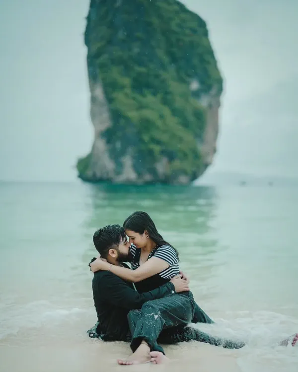 An intimate embrace in the shallow waters of a secluded Thai beach, with a dramatic rock formation in the background.