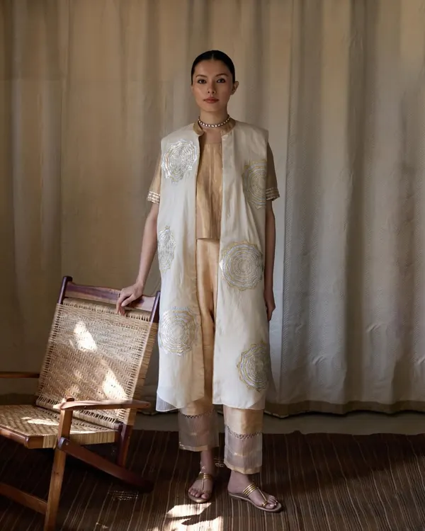 A full-length shot for Bellerobe, showcasing a layered ethnic ensemble. The natural light from the window highlights the delicate embroidery on the off-white jacket, while the simple cane chair adds a touch of organic texture to the scene.